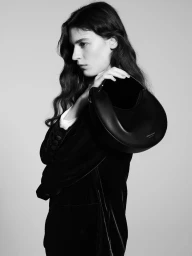 Model with long dark hair wearing a black jacket holding a curved leather handbag against a plain grey backdrop.