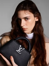 Close-up portrait of a woman holding a black leather handbag with a silver logo against a plain grey background
