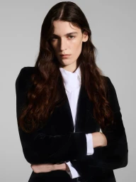 Model with long dark wavy hair wearing a black jacket over a white collared shirt against a grey studio backdrop