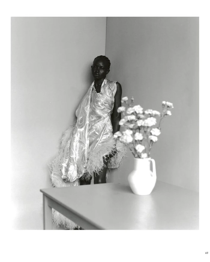 Model Aweng Chuol in a feathered dress leaning in a room corner beside a table with a white vase of flowers.