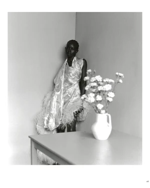 Model Aweng Chuol in a feathered dress leaning in a room corner beside a table with a white vase of flowers.