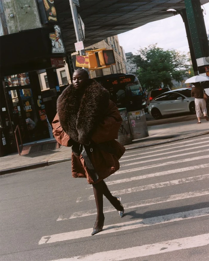 Model Aweng Chuol in a brown puffer coat with a large fur collar walking across a city street.