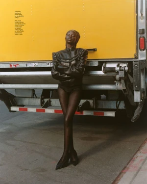 Aweng Chuol posing against a yellow truck in black leather jacket and headwrap for Heroine Magazine
