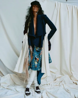 Model wearing a dark blue jacket, patterned skirt, hat, and white leg warmers against a draped fabric background.