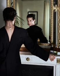 Man wearing a black suit jacket leaning on a white fireplace mantel, looking into an ornate gilded mirror.
