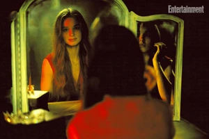 A young woman with long hair seated in front of a three-paneled vanity mirror reflecting her image under dramatic red and green lighting.