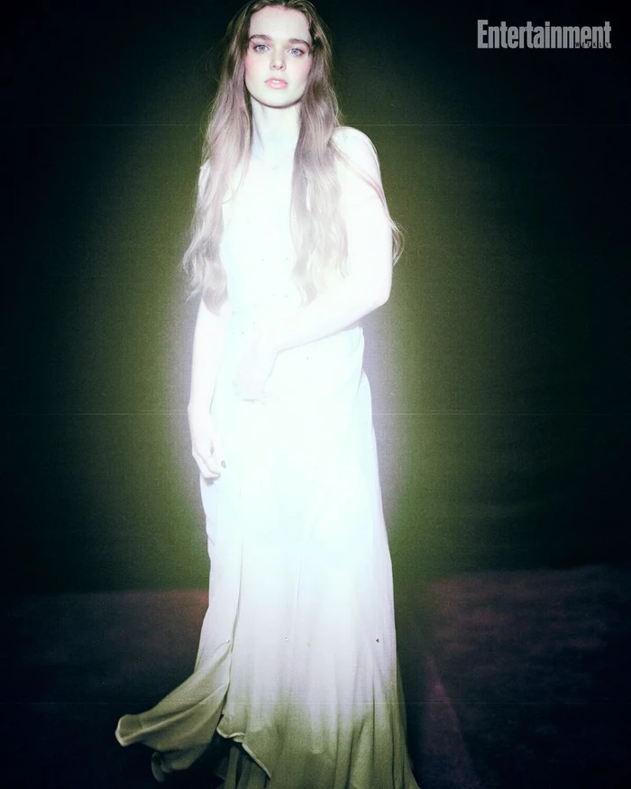 A woman in a long white dress standing against a dark background with an Entertainment Weekly watermark in the upper right corner.