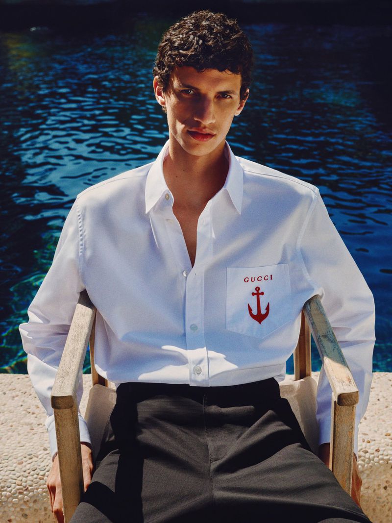 Man in white shirt with anchor logo sitting in chair by pool. Dark hair, medium shot in outdoor setting with blue background.