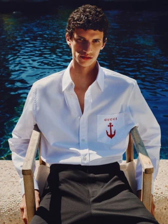 Man in white shirt with anchor logo sitting in chair by pool. Dark hair, medium shot in outdoor setting with blue background.