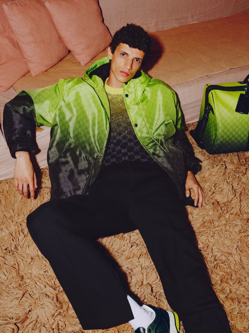 An adult male model poses on a carpet with a bright green and black jacket and a matching duffel bag in a fashion shot.