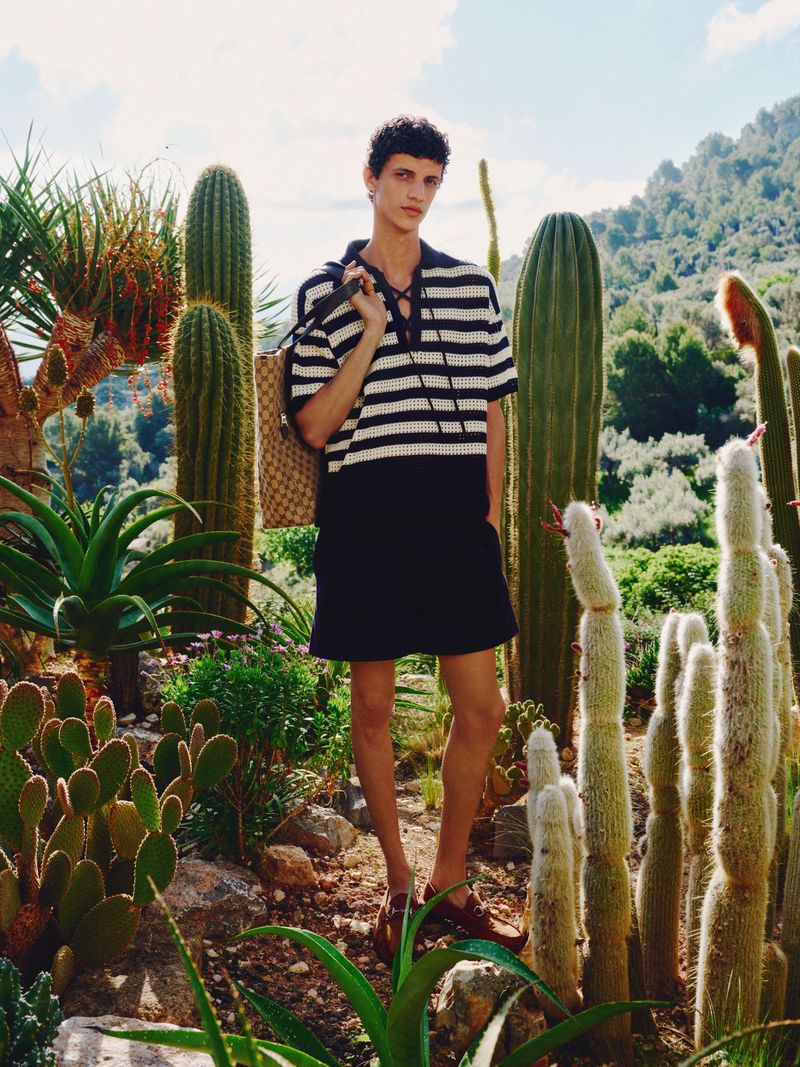 Man wearing striped shirt and black shorts poses in desert garden setting surrounded by cactus and lush green desert foliage, sunny day.