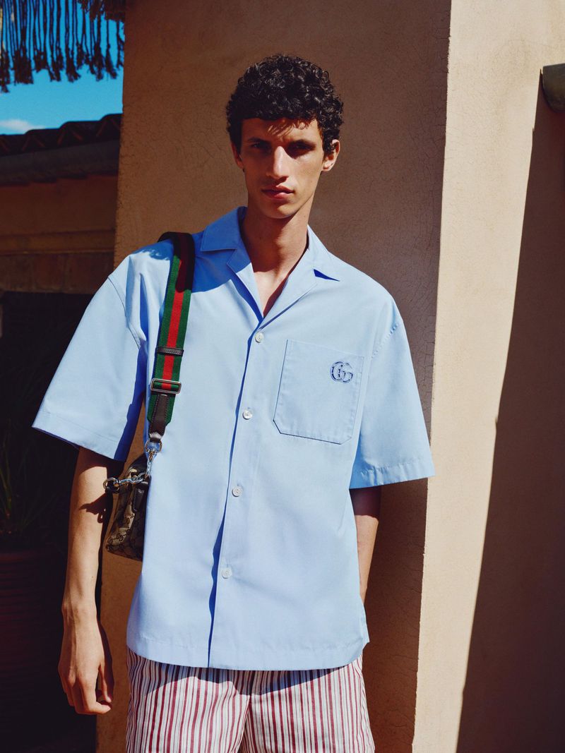 A young male model poses outdoors in a light blue collared shirt, red and white striped shorts, and a crossbody bag with a red and green strap.