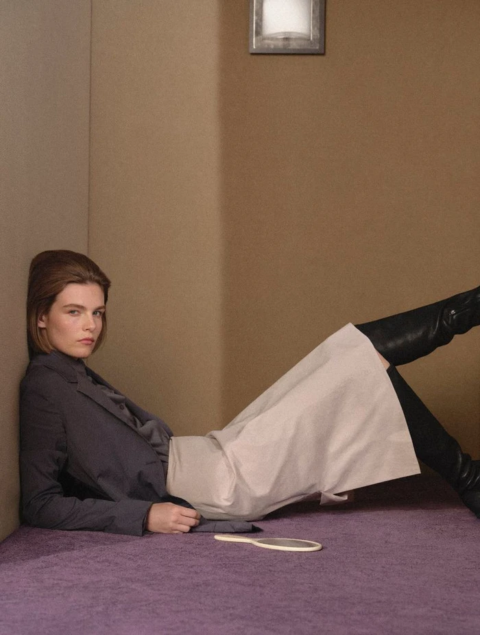 Model reclining on a purple carpeted floor in a dark jacket and skirt with high black boots for CR Fashion Book.
