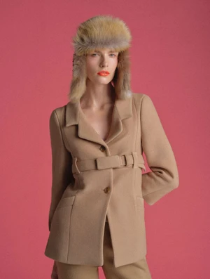 Model wearing a tan belted jacket and furry winter hat against a solid pink background