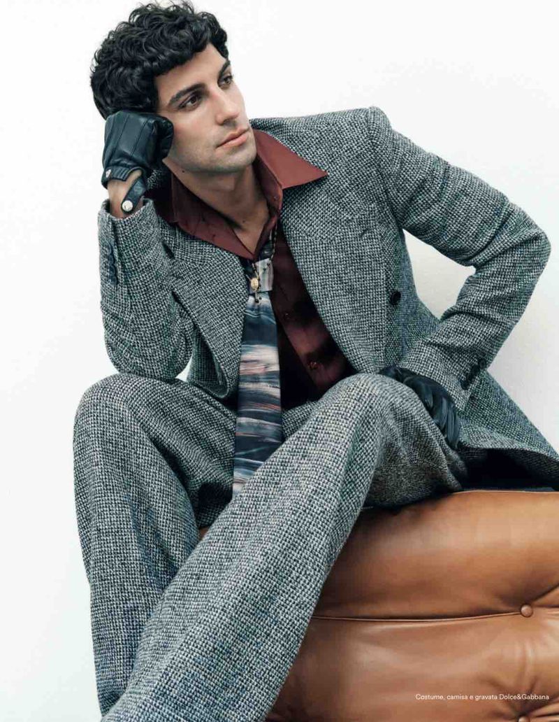 Model Francisco Faria seated on a leather ottoman wearing a tweed suit, red shirt, and black leather gloves.