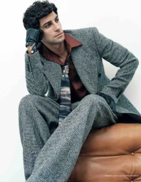 Model Francisco Faria seated on a leather ottoman wearing a tweed suit, red shirt, and black leather gloves.