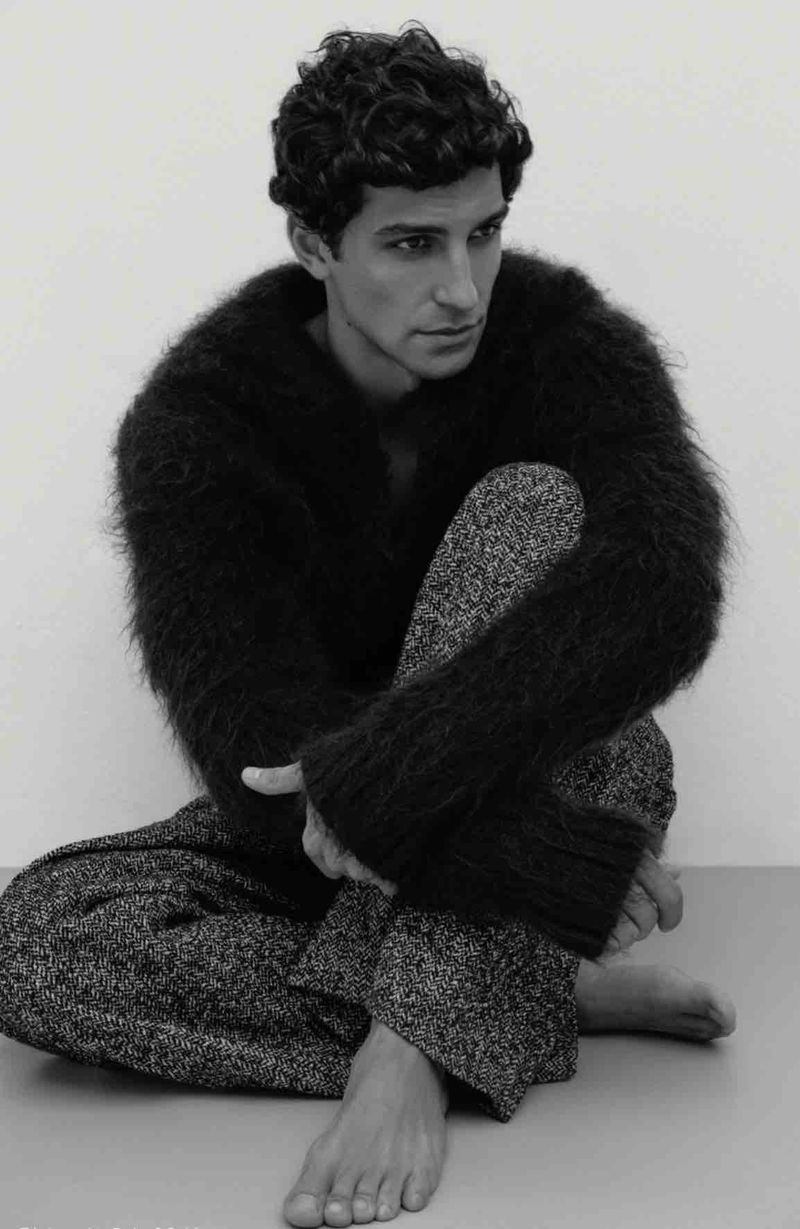 Model Francisco Faria seated on the floor wearing a dark fuzzy sweater and patterned trousers in a studio setting.