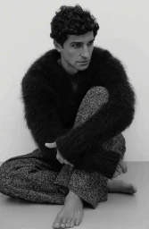 Model Francisco Faria seated on the floor wearing a dark fuzzy sweater and patterned trousers in a studio setting.