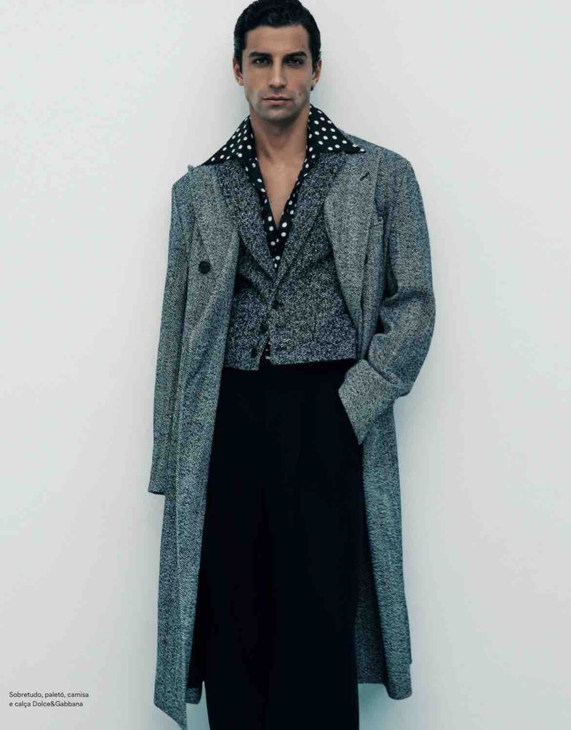 Man wearing a grey tweed coat, patterned vest, and dark trousers against a plain white studio background.
