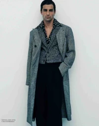 Man wearing a grey tweed coat, patterned vest, and dark trousers against a plain white studio background.