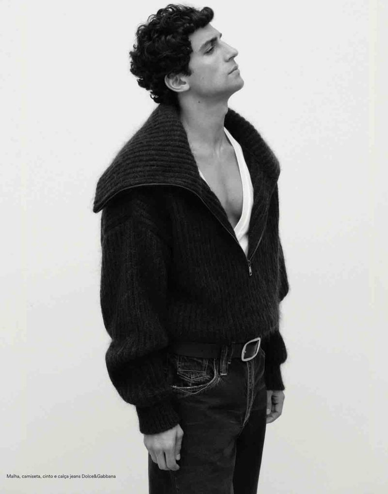 Man with curly hair wearing a dark zip sweater and jeans against a white seamless background