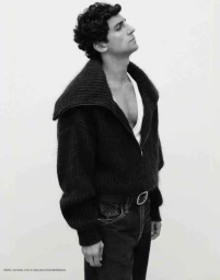 Man with curly hair wearing a dark zip sweater and jeans against a white seamless background