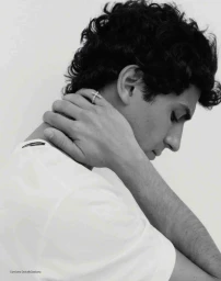 Profile portrait of a male model with dark curly hair wearing a white t-shirt, hand on neck against a light background.
