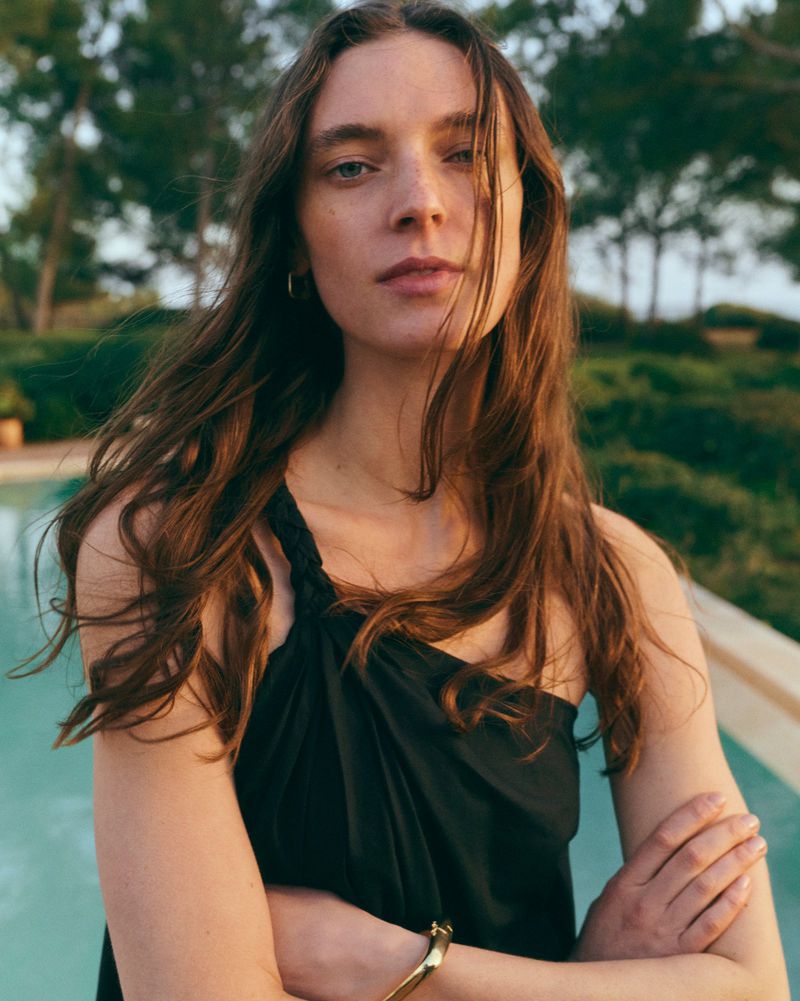 Adult woman with long brown hair poses in a sleeveless black top against an outdoor setting with trees and a turquoise pool.