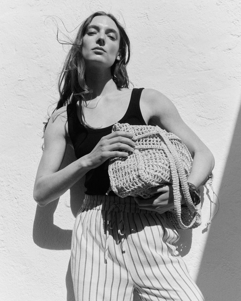 Monochrome image of a woman in a tank top and striped pants, holding a knitted bag against a textured white wall with sunlight.