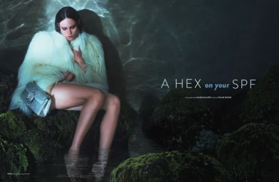 Model wearing a fluffy white coat and holding a silver bag seated on mossy rocks near dark water for Flaunt project A Hex on Your SPF