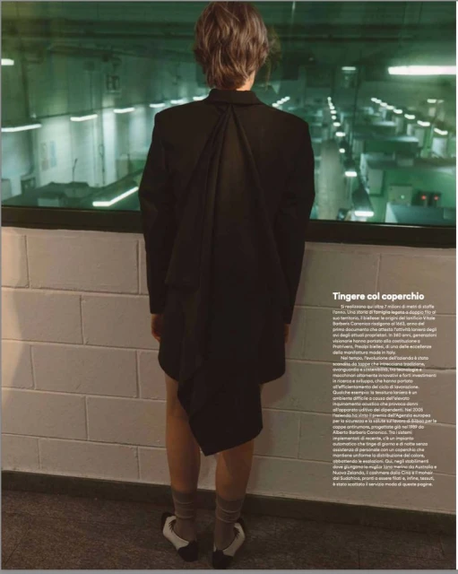 Model seen from behind wearing a black blazer and patterned socks against a white block wall and industrial background