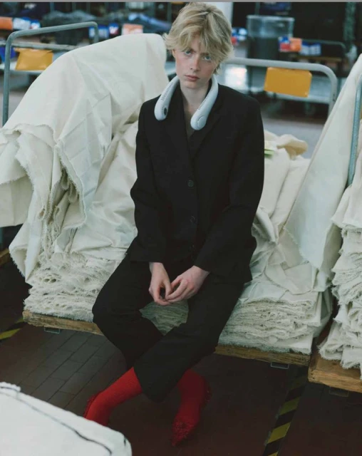 Model in black suit and red socks seated amongst stacked white fabric rolls in a warehouse