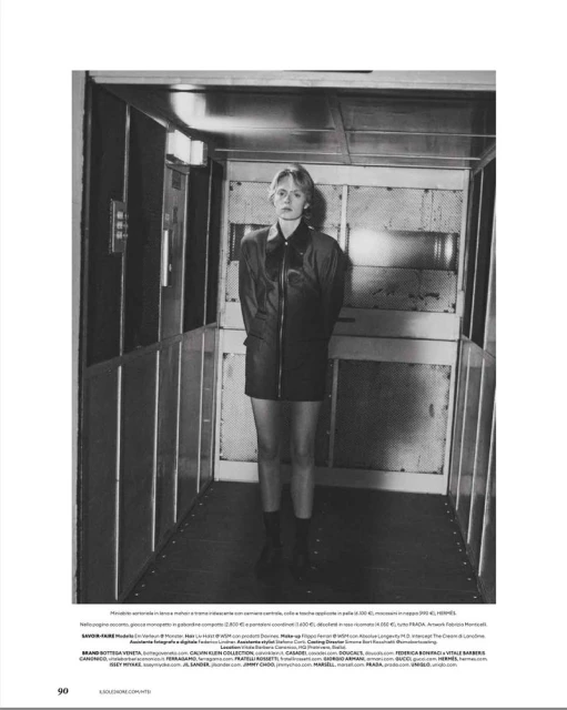 Model standing inside an elevator cabin wearing a dark leather coat and dark shoes in a black and white editorial layout.