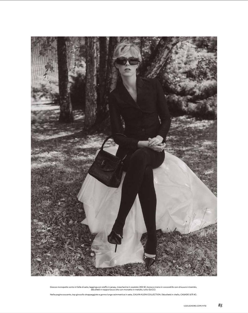 Model in dark sunglasses and black blazer seated on a white draped structure outdoors amidst trees