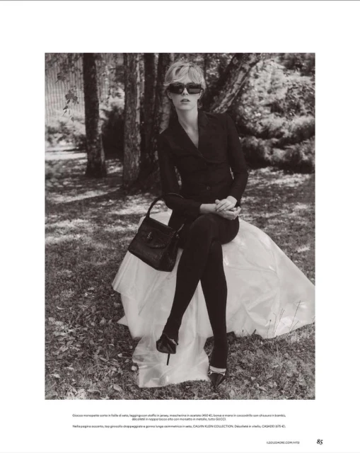 Model in dark sunglasses and black blazer seated on a white draped structure outdoors amidst trees