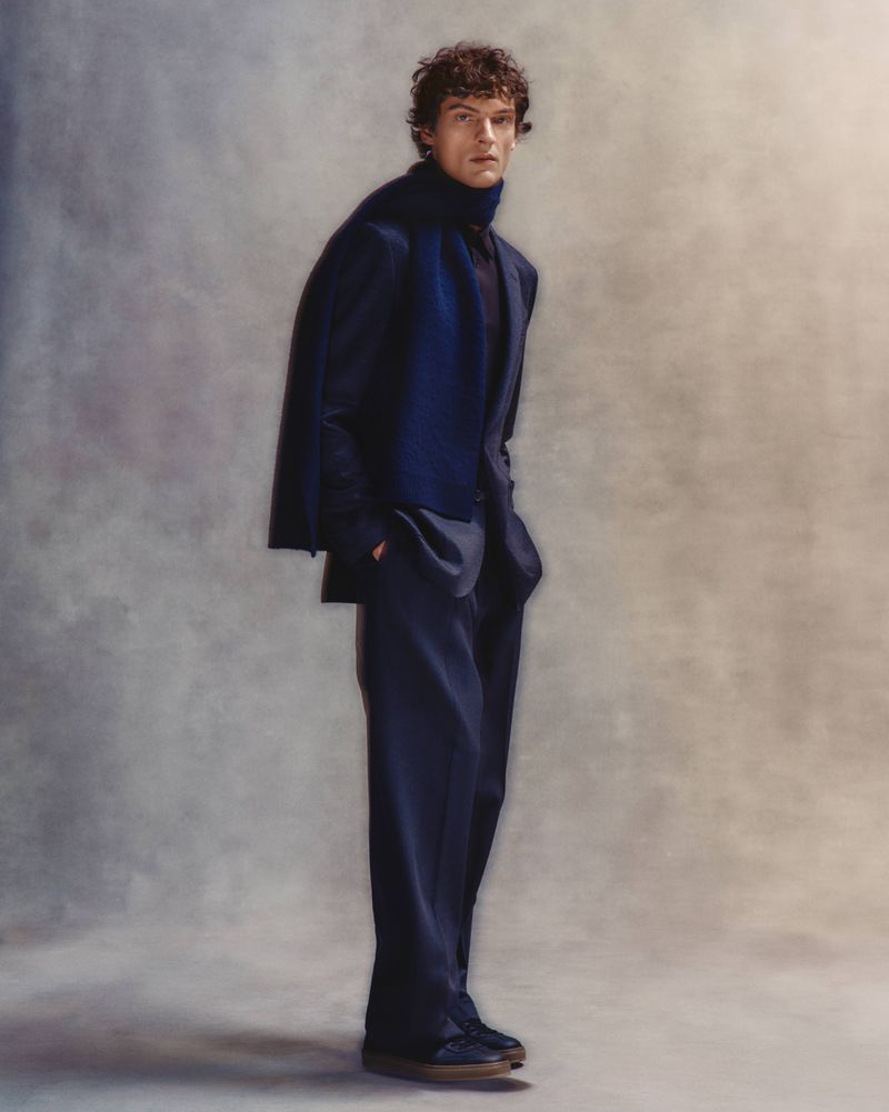 Model in navy suit and matching scarf posing against a textured grey studio backdrop for BOSS by David Beckham.
