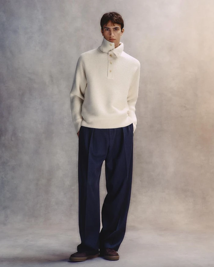 Model standing against a mottled textured backdrop wearing a white high-neck sweater and dark trousers
