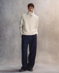 Model standing against a mottled textured backdrop wearing a white high-neck sweater and dark trousers