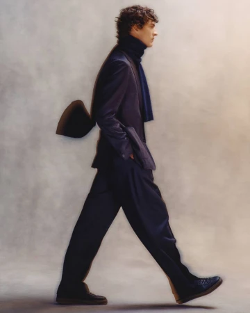 Model in a dark suit and turtleneck walking against a neutral textured studio backdrop
