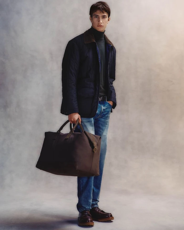 Model standing in a quilted jacket and blue jeans holding a large dark duffel bag against a textured grey background.