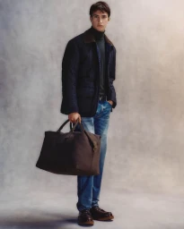 Model standing in a quilted jacket and blue jeans holding a large dark duffel bag against a textured grey background.