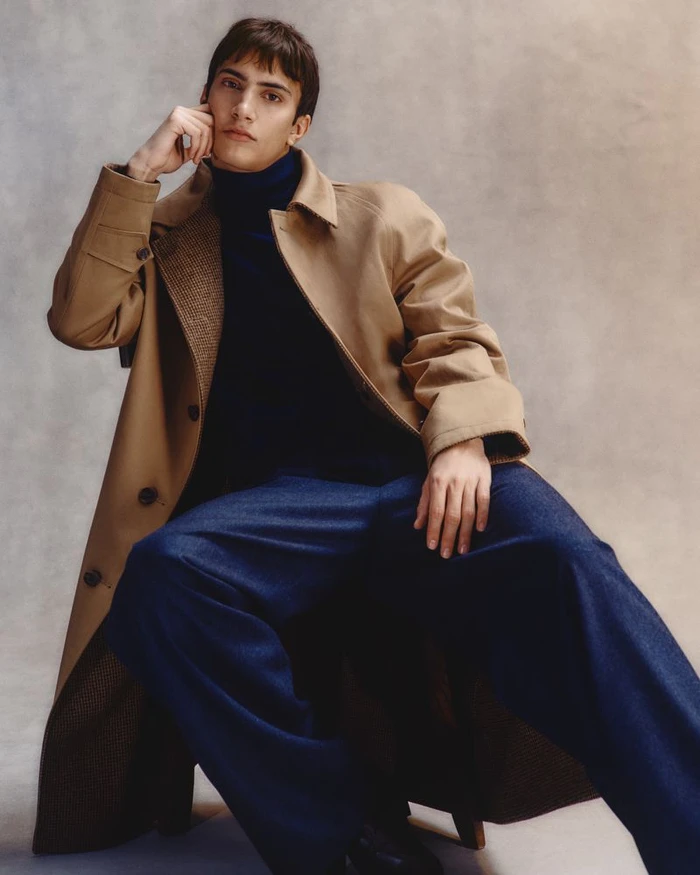 Model seated wearing a camel coat and dark blue trousers against a textured studio background.
