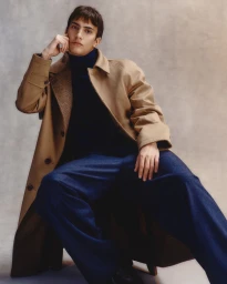 Model seated wearing a camel coat and dark blue trousers against a textured studio background.