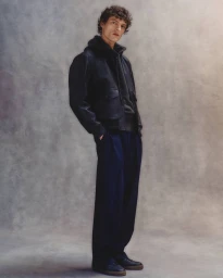 Model in dark leather jacket and navy trousers standing against a textured backdrop