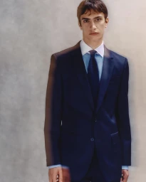A young male model in a dark navy suit with a white shirt and blue tie standing against a textured studio background.
