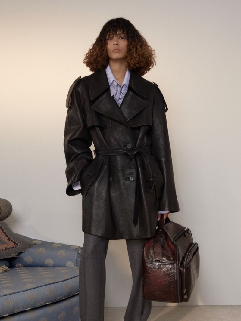 Model wearing a black leather trench coat over a light shirt and holding a large dark leather travel bag in a studio setting