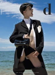 Model wearing a black coat, white shirt, and leather cap standing outdoors with an ocean and clear sky background.