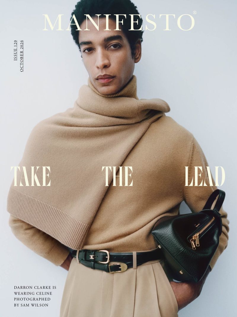 Male model wearing a camel turtleneck sweater and trousers holding a dark green handbag against a white background.