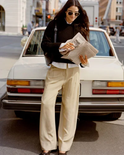 Model in sunglasses and black sweater leaning against a white car holding a newspaper on a city street