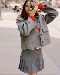 Model holding a vintage camera wearing a grey wool jacket and pleated skirt on a city street